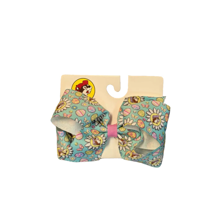 Pastel Teal Buc-ee’s Medium Easter hair bow. The vibrant teal ribbon is decorated with spring daisies and multicolored Easter eggs and the Buc-ee’s beaver logo wearing a purple hat.. The center of the bow is tied with a contrasting pink fabric knot, and it is mounted on a white "Buc-ee’s" branded display card. Displayed on a clean white background.