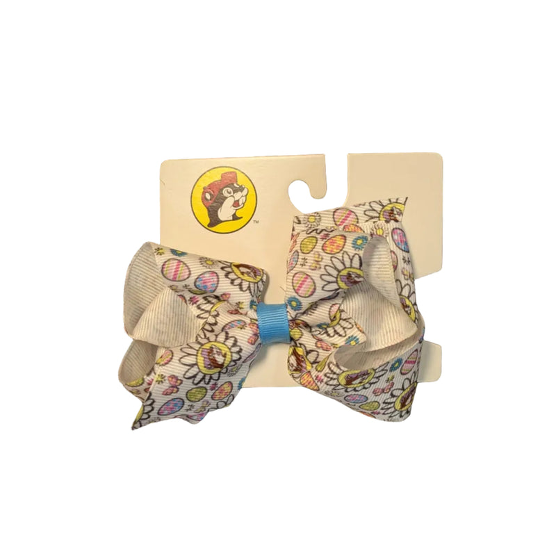 Classic White Medium sized Buc-ee’s Easter hair bow. The bow is made of textured ribbon with a white background covered in colorful floral and egg patterns and the Buc-ee’s beaver logo wearing a purple hat. The center is secured with a small blue fabric knot, and the retail packaging features the red-hatted Buc-ee's logo at the bottom. Displayed on a clean white background.