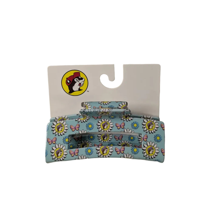 A flat-lay photograph of a Buc-ee’s branded hair claw clip on a white retail display card, arranged against a plain white background. The cliip is a rectangular, open-frame claw clip with a vibrant teal base, featuring a pattern of white daisies, colorful butterflies, and the Buc-ee's beaver logo with a purple hat.