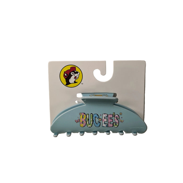 A flat-lay photograph of a Buc-ee’s branded hair claw clip on a white retail display cards, arranged against a plain white background. A solid, curved light blue claw clip featuring the word "BUC-EE'S" in a playful, multi-colored bubble font. 