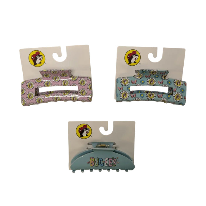 A flat-lay photograph of three Buc-ee’s branded hair claw clips on their white retail display cards, arranged against a plain white background. A pink clip and teal clip on the top featuring all over butterfly and white daisy print with the Buc-ee beaver logo with a purple hat. Bottom clip is solid teal with the word Buc-ee's in multi-colored bubble font.