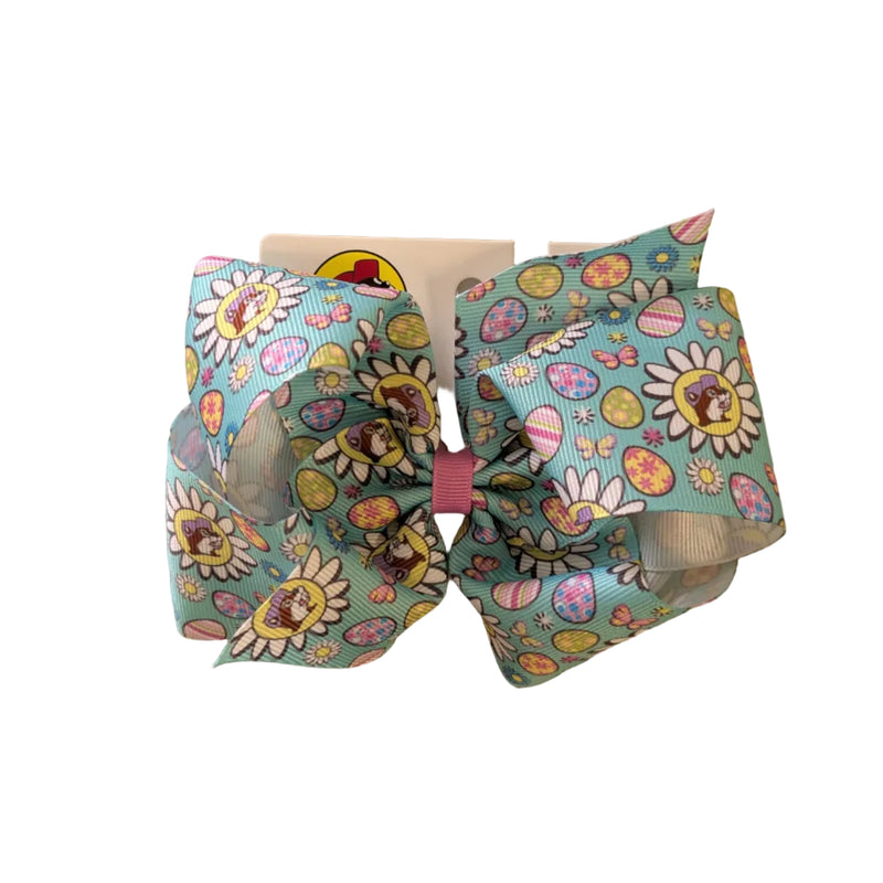A Large Pastel Teal Buc-ee’s Easter hair bow. The vibrant teal ribbon is decorated with spring daisies and multicolored Easter eggs and the Buc-ee’s beaver logo wearing a purple hat. The center of the bow is tied with a contrasting pink fabric knot, and it is mounted on a white "Buc-ee’s" branded display card. Displayed on a clean white background.