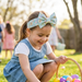 A bright and joyful lifestyle photograph of a young girl participating in an outdoor Easter egg hunt on a sunny day. She is wearing a denim jumper and the teal Buc-ee's Easter headband with a large statement bow. She is crouching in the green grass, reaching for a bright pink plastic egg while holding a wicker basket filled with colorful treats. The background is softly blurred, showing other children playing in a sun-drenched yard, creating a warm and festive holiday atmosphere.