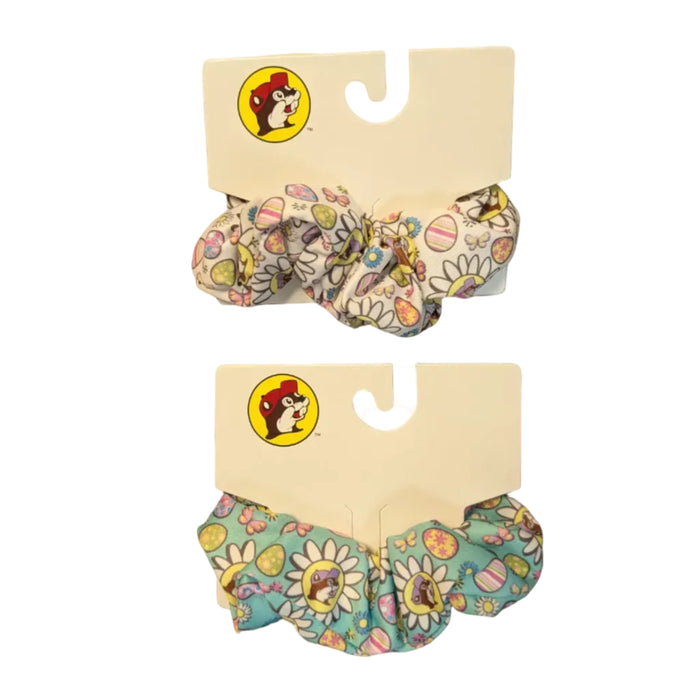 Two Buc-ee’s Easter-themed hair scrunchies are displayed on their original white cardboard hanging tags against a white background. The top scrunchie has a white background, while the bottom scrunchie has a light teal background. Both feature a repeating festive pattern of colorful Easter eggs, butterflies, and white daisies with the Buc-ee’s beaver mascot, wearing a purple hat, centered inside the flower petals.