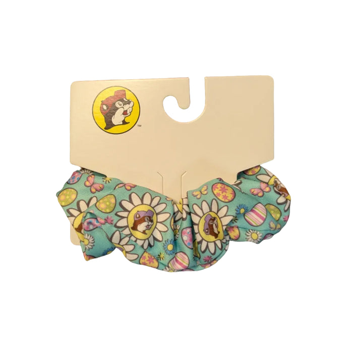 A close-up of a light teal (blue) Buc-ee’s Easter hair scrunchie shown on its white cardboard hanging tag. The fabric features a repeating festive pattern of colorful Easter eggs, small butterflies, and white daisies. The iconic Buc-ee’s beaver mascot is prominently featured in the center of the daisies, wearing a purple hat. Scrunchie displayed on a clean white background.