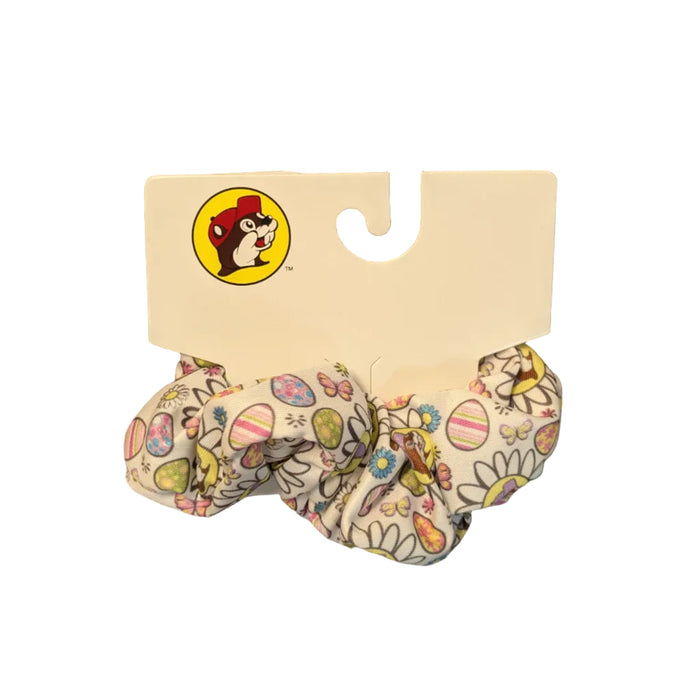 A close-up shot of an official Buc-ee’s Easter hair scrunchie on its white cardboard hanging tag. The scrunchie features a white fabric background with a festive spring pattern consisting of pastel Easter eggs, colorful butterflies, and white daisies. The Buc-ee’s beaver mascot, wearing a purple hat, is centered inside the larger daisies throughout the design. Scrunchie is displayed on a clean white background.