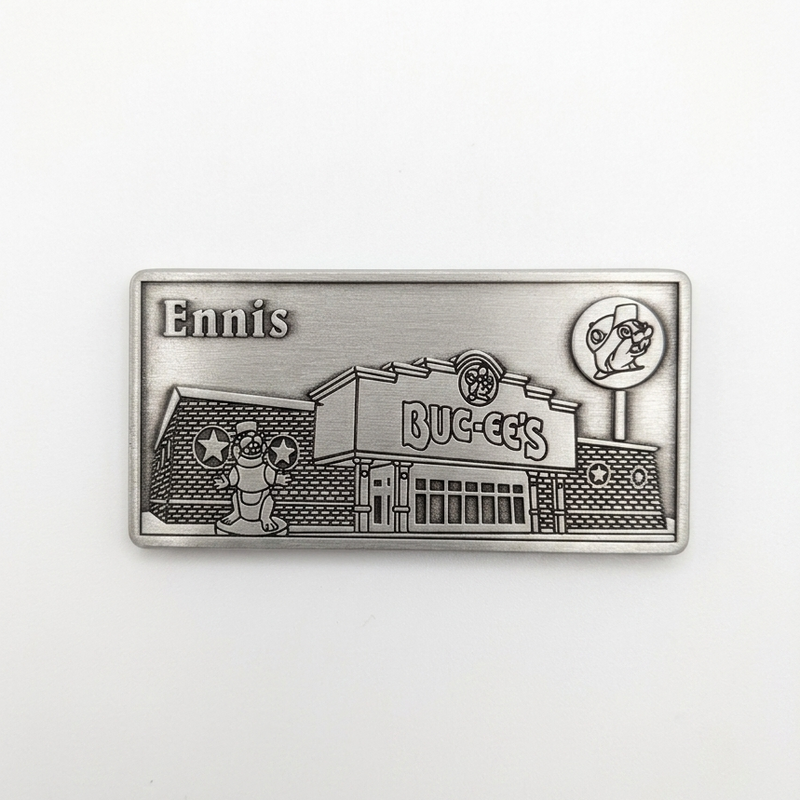 A rectangular pewter metal magnet showing an embossed 3D relief of the Buc-ee's Ennis travel center building, complete with the beaver statue out front, the Buc-ee's sign, a lollipop road sign in the upper right, and the word "Ennis" in the upper left corner.