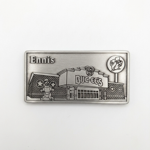 A rectangular pewter metal magnet showing an embossed 3D relief of the Buc-ee's Ennis travel center building, complete with the beaver statue out front, the Buc-ee's sign, a lollipop road sign in the upper right, and the word "Ennis" in the upper left corner.