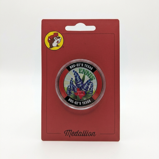 A circular metal souvenir Buc-ee's Ennis, TX medallion coin displayed inside a clear acrylic protective capsule on a red card. The coin face features bluebonnets, a red Texas outline, "ENNIS" written across the top, and a black rim stamped with "BUC-EE'S TEXAS".