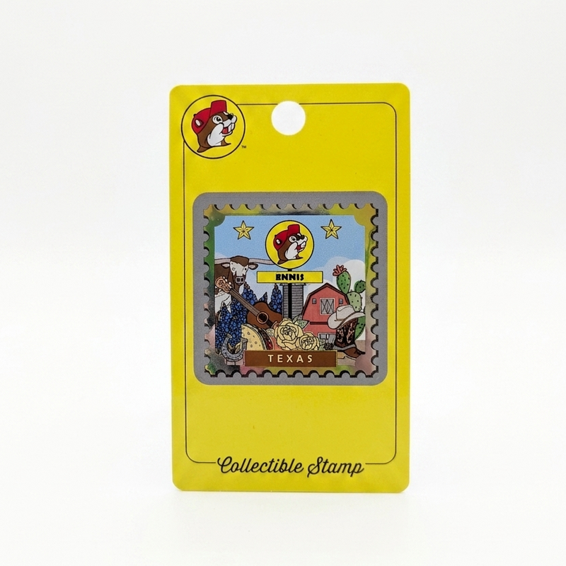 A postage-stamp-style souvenir with perforated metallic silver edges on a yellow packaging card. The stamp artwork includes a Buc-ee's Ennis sign, Texas Longhorn, guitar, red barn, bluebonnets, and cowboy boot, with "TEXAS" printed at the bottom.