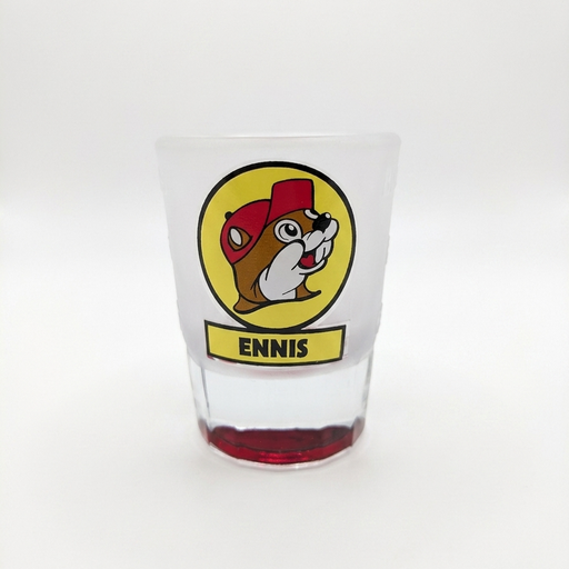 A tapered, frosted clear glass shot glass featuring the yellow circular Buc-ee's beaver logo with a yellow banner reading "ENNIS" underneath. The thick, solid glass base is tinted in a translucent red color.