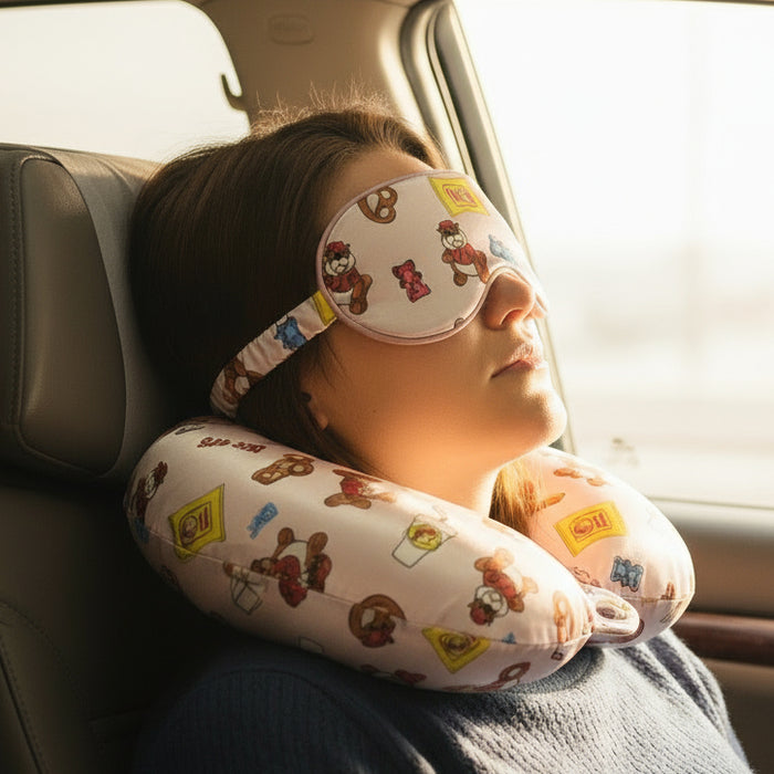 Buc-ee’s travel set featuring a silky eye mask and matching U-shaped neck pillow decorated with Buc-ee the Beaver, Beaver Nuggets bags, gummy bears, pretzels, and snack icons, displayed on a woman sitting in a car.