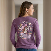 Woman indoors wearing the heathered purple Buc-ee's long-sleeve t-shirt, showcasing the back graphic of Buc-ee inside a floral wreath with the banner "Find Joy in the Journey."