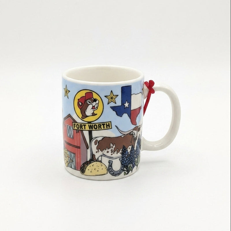 A white ceramic mini mug against a clean white background featuring a light blue sky wrapped in Texas-themed illustrations. The graphics include a red barn, a Texas longhorn steer, a taco, bluebonnets, a horseshoe, a Texas flag map outline, and a yellow Buc-ee's sign reading "FORT WORTH". A small red hanging string is tied to the handle.