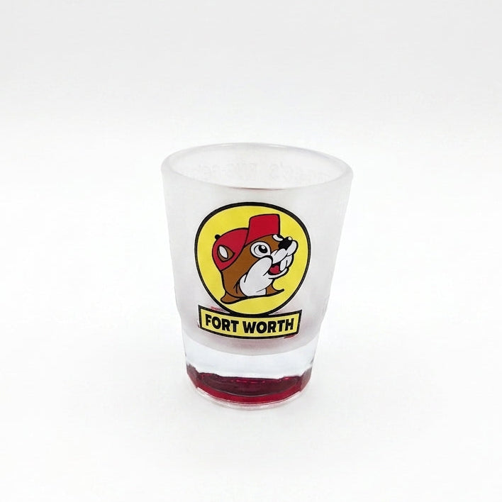 A tapered, frosted clear glass shot glass featuring the iconic yellow circular Buc-ee's beaver logo with a yellow banner reading "FORT WORTH" underneath. The heavy, solid glass base is tinted in a vibrant, translucent red color.