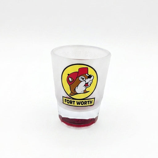 A tapered, frosted clear glass shot glass featuring the iconic yellow circular Buc-ee's beaver logo with a yellow banner reading "FORT WORTH" underneath. The heavy, solid glass base is tinted in a vibrant, translucent red color.