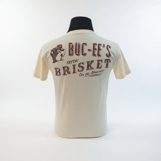 Back view of cream-colored Buc-ee’s T-shirt showing “Buc-ee’s Fresh Brisket on the Board!!” text and beaver logo in red print, displayed on a man standing in front of a grill in a backyard. 