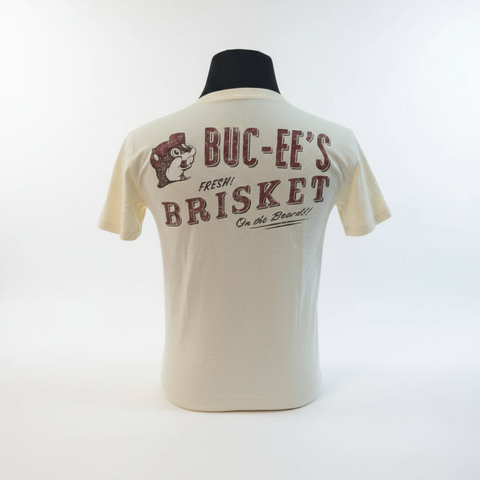 Back view of cream-colored Buc-ee’s T-shirt showing “Buc-ee’s Fresh Brisket on the Board!!” text and beaver logo in red print, displayed on a man standing in front of a grill in a backyard. 