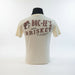 Back view of cream-colored Buc-ee’s T-shirt showing “Buc-ee’s Fresh Brisket on the Board!!” text and beaver logo in red print, displayed on a man standing in front of a grill in a backyard. 