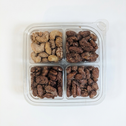 A top-down product shot of a 4-compartment clear plastic tray against a clean white background. The tray features four different varieties of glazed nuts: pecans, walnuts, almonds, and cashews, each separated into its own square section. The clear plastic packaging is visible with a tab in the upper-right corner for opening.