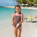 A girl standing on a sandy beach wearing a Buc-ee's one-piece leopard print swimsuit with pink straps. Pink Buc-ee beaver logos are scatterd throughout the leopard print on the swimsuit.