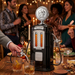 A lifestyle photo of a retro-style beverage dispenser shaped like a gas pump, set in a party environment. The dispenser has a circular top with a beaver mascot logo and two vertical glass tubes filled with amber liquid, marked with numbers 1 through 8. A person’s hand on the left is using a silver fuel-nozzle dispenser to pour a drink into an ice-filled glass, while a second nozzle rests in its holster on the right side of the unit. A front plate on the dispenser is embossed with the words HIGH OCTANE.