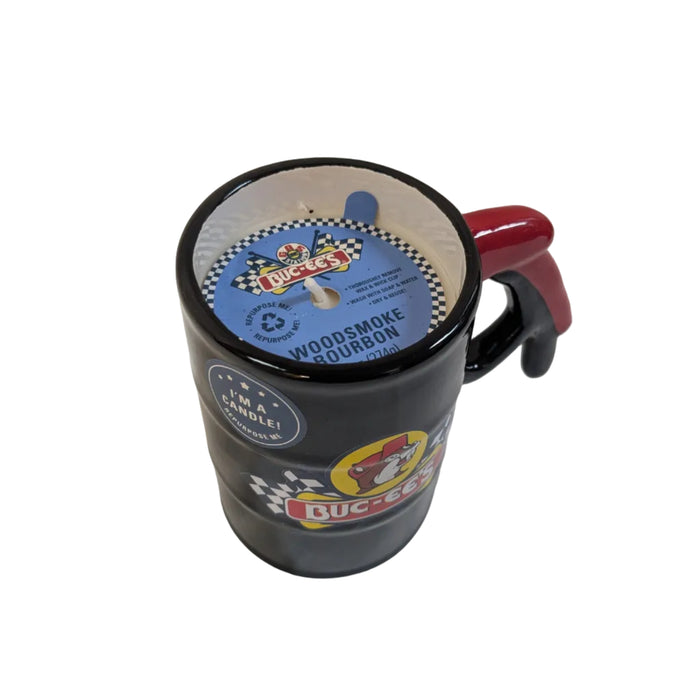An overhead, angled view of the black ceramic Buc-ee’s gas pump handle candle. A blue paper insert sits on top of the white wax, displaying the scent "WOODSMOKE BOURBON" and instructions to wash and reuse the container once finished. A small blue circular sticker on the side reads, "I'M A CANDLE! REPURPOSE ME." Displayed on a clean white background.