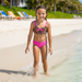 A young girl smiling on a beach wearing a Buc-ee's Girls Leopard Two Piece Swimsuit. A leopard print bikini top with a pink bow and pink straps. Solid pink bottoms with a pink Buc-ee beaver logo on the top left side of the swim bottoms.