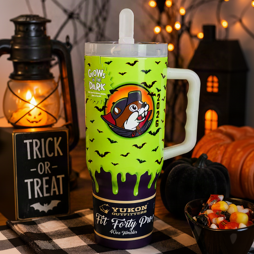 A Halloween-themed insulated tumbler with a handle, featuring a bright lime green body with black bat silhouettes, dripping dark green slime, a cartoon dog mascot in an orange circle, and a dark purple band at the bottom. The tumbler is set on a black-and-white checkered tablecloth with a lit lantern, pumpkin, and string lights in the background.