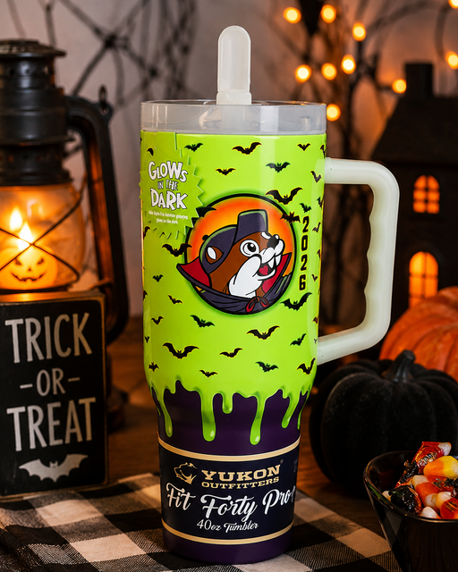 A Halloween-themed insulated tumbler with a handle, featuring a bright lime green body with black bat silhouettes, dripping dark green slime, a cartoon dog mascot in an orange circle, and a dark purple band at the bottom. The tumbler is set on a black-and-white checkered tablecloth with a lit lantern, pumpkin, and string lights in the background.