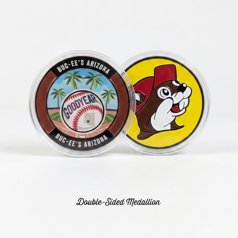 A studio shot showcasing both sides of a Buc-ee's Goodyear, Arizona collectible metal medallion side-by-side inside clear protective cases against a plain white background. The left side displays a colorful baseball and palm tree design reading "GOODYEAR" and "BUC-EE'S ARIZONA." The right side displays the classic yellow and red Buc-ee's beaver mascot logo. Elegant cursive text at the bottom reads "Double-Sided Medallion".