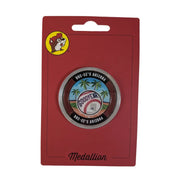 A photo of a circular metal Buc-ee's Goodyear, Arizona medallion on its red cardboard packaging. The top-left corner features the small, round Buc-ee's beaver logo, and the word "Medallion" is written in cursive script at the bottom. Inside a clear protective plastic casing, the double-sided coin shows a baseball design with the word "GOODYEAR" on it, set against a background of green grass, palm trees, and a blue sky. The thick dark brown outer border of the coin reads "BUC-EE'S ARIZONA" on top and bottom.