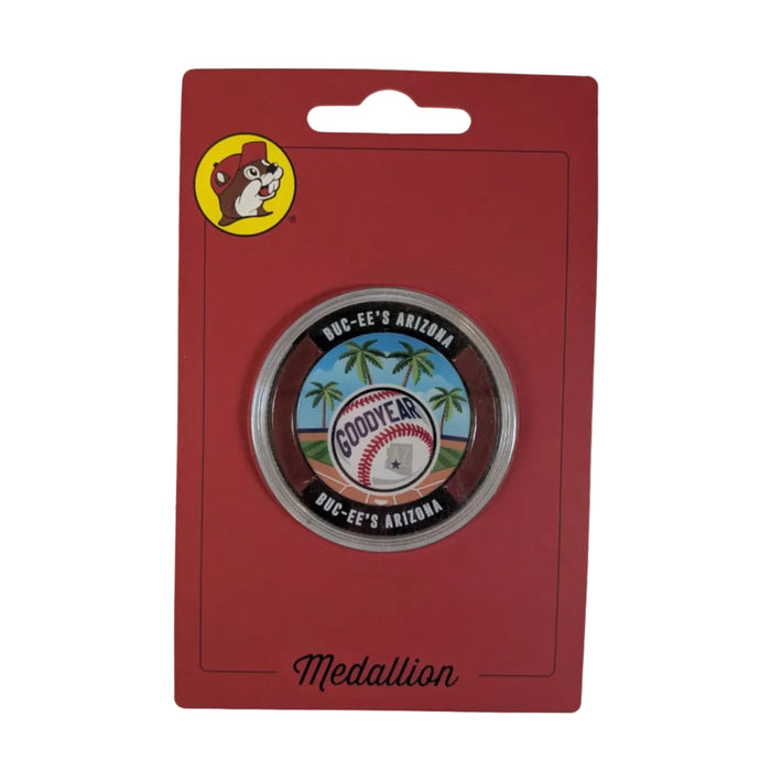 A photo of a circular metal Buc-ee's Goodyear, Arizona medallion on its red cardboard packaging. The top-left corner features the small, round Buc-ee's beaver logo, and the word "Medallion" is written in cursive script at the bottom. Inside a clear protective plastic casing, the double-sided coin shows a baseball design with the word "GOODYEAR" on it, set against a background of green grass, palm trees, and a blue sky. The thick dark brown outer border of the coin reads "BUC-EE'S ARIZONA" on top and bottom.