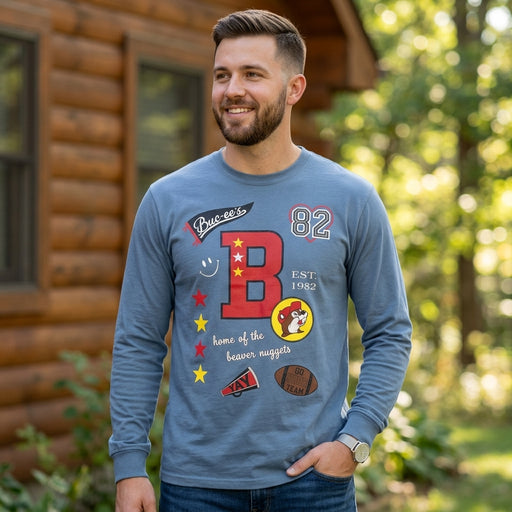 Man wearing the Buc-ee's slate blue varsity graffiti long-sleeve shirt outdoors, featuring a front collage graphic with a large red letter B, Buc-ee's logo badge, and sports accents.