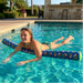 A lifestyle image of a smiling young woman floating in an outdoor swimming pool, resting her arms across a large blue and green Buc-ee's premium pool noodle. The pool noodle features an all-over repeating pattern of circular Buc-ee's beaver logos where the mascot wears a bright green cap, perfectly matching the solid green circular cap on the end of the noodle. Clear, sunlit pool water ripples around her, with a blurred resort-style patio, palm trees, and an outdoor cabana bar is visible.
