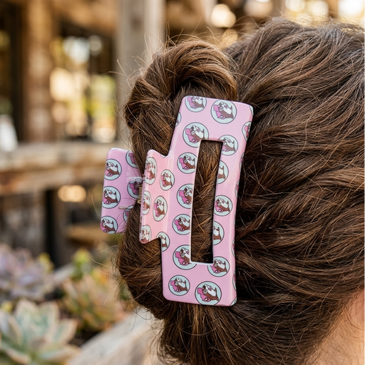 A lifestyle close-up from behind a woman with wavy brunette hair partially tied back in a bun. Secured in her hair is a pink rectangular claw clip with a repeating pattern of the Buc-ee’s beaver mascot inside small blue circles. The setting is a bright, sunlit outdoor patio with blurred succulents and wooden furniture in the background.