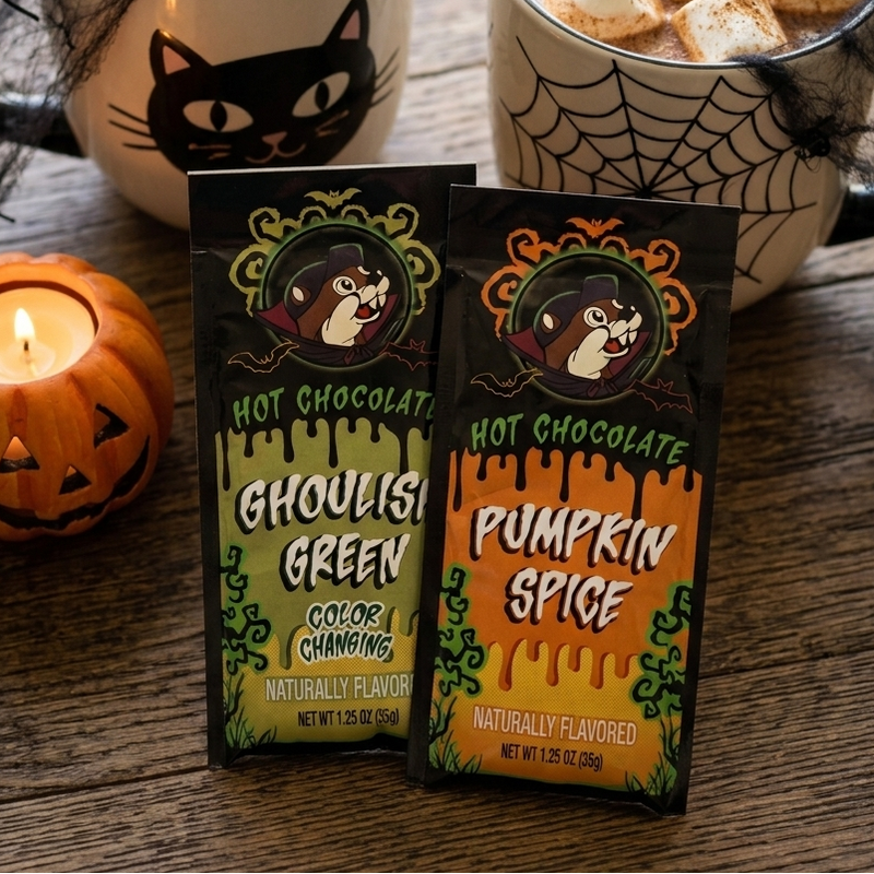 A lifestyle photograph on a rustic wooden surface featuring two Buc-ee's Halloween hot chocolate packets—"Ghoulish Green Color Changing" and "Pumpkin Spice"—lying flat in the foreground. Behind them are two festive Halloween mugs filled with hot cocoa and marshmallows, alongside a glowing jack-o'-lantern candle.