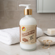 A close-up lifestyle shot of a frosted glass bottle of Buc-ee's Glazed Pecans Hand & Body Lotion sitting on a marble bathroom counter. A neatly rolled white towel and a small gold jewelry dish are visible in the background against a light neutral tiled wall.
