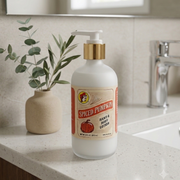 A frosted glass bottle of Buc-ee's "Spiced Pumpkin" hand and body lotion with a gold pump sits on a bathroom counter. A small vase with eucalyptus and olive branches is to the left, and a chrome faucet is in the background.