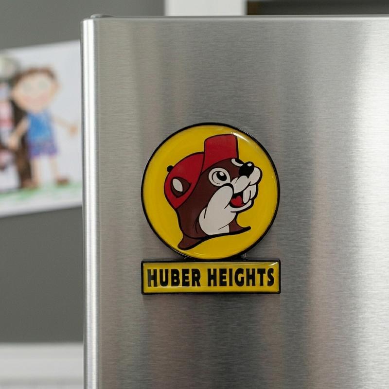 A close-up, slightly angled lifestyle photograph of a Buc-ee’s souvenir refrigerator magnet attached to a brushed stainless steel fridge. The magnet features the iconic Buc-ee’s beaver mascot wearing a red cap on a circular yellow background, with a rectangular yellow bar below it reading "HUBER HEIGHTS" in bold black letters. The background is softly blurred, showing the corner of the refrigerator and a hint of a child's drawing on the wall.