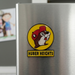 A close-up, slightly angled lifestyle photograph of a Buc-ee’s souvenir refrigerator magnet attached to a brushed stainless steel fridge. The magnet features the iconic Buc-ee’s beaver mascot wearing a red cap on a circular yellow background, with a rectangular yellow bar below it reading "HUBER HEIGHTS" in bold black letters. The background is softly blurred, showing the corner of the refrigerator and a hint of a child's drawing on the wall.