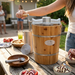 A lifestyle photo of a woman using a wooden bucket ice cream maker on a backyard picnic table during a summer gathering. Nearby, a person holds a scoop of ice cream, and two bowls of freshly served ice cream with toppings sit on the table.