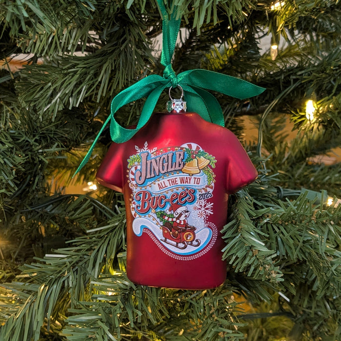 Back view of red T-shirt-shaped Buc-ee’s 2025 Christmas ornament on a tree, showing the festive “Jingle All the Way to Buc-ee’s” design with a holiday truck and candy cane details.