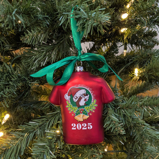 Red T-shirt-shaped Buc-ee’s 2025 Christmas ornament hanging on a Christmas tree with green ribbon and lights, featuring Buc-ee wearing a Santa hat above holly and bells.