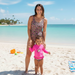 A smiling woman and a young toddler pose together on a sun-drenched beach with turquoise water and palm trees in the background. The woman is wearing a Buc-ee's one-piece leopard print swimsuit featuring small pink details. The toddler stands in front of her, wearing a bright pink long-sleeve rash guard with a central circular Buc-ee logo and matching leopard print swim bottoms.