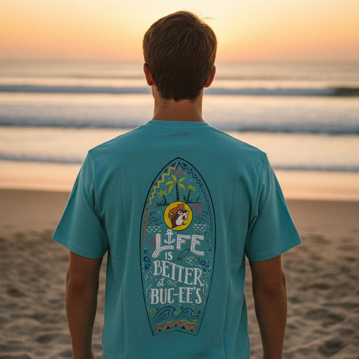 A man standing on the beach showing the back view of the 'Life is Better at Buc-ee's' Turquoise Shirt showing a large surfboard graphic with palm trees, waves, and the text 'Life is Better at Buc-ee's'."