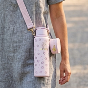 A woman walking along a paved path in a park. She is wearing a grey linen short-sleeved dress and has a light pink Buc-ee's floral hands-free tumbler worn across her shoulder with a matching pink strap. The light pink water bottle features a pattern of small pink roses with green leaves and a prominent gold Buc-ee's logo in the center. A small, matching pink zippered pouch is attached to the side of the tumbler. 