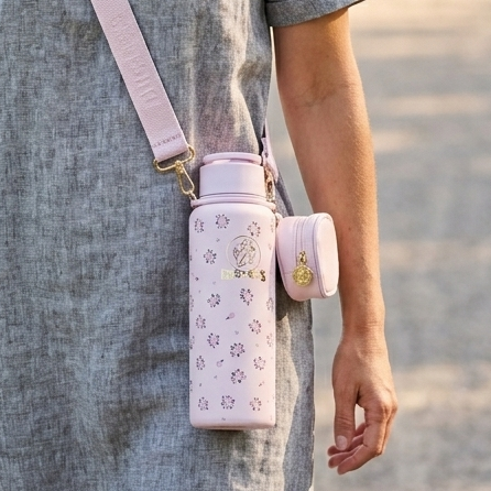 A woman walking along a paved path in a park. She is wearing a grey linen short-sleeved dress and has a light pink Buc-ee's floral hands-free tumbler worn across her shoulder with a matching pink strap. The light pink water bottle features a pattern of small pink roses with green leaves and a prominent gold Buc-ee's logo in the center. A small, matching pink zippered pouch is attached to the side of the tumbler. 