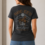 Woman shown from behind wearing a dark obsidian-gray Buc-ee’s T-shirt with a vintage Western “BUC-EE’S Local Legend” cowboy and horse graphic on the back, paired with blue jeans in a neutral indoor setting.