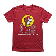 A flat lay product photograph of a red Buc-ee’s t-shirt centered on a clean, solid white background. The shirt features a large yellow circular logo of the Buc-ee's beaver mascot wearing a red hat, with "BUC-EE'S" and "HUBER HEIGHTS, OH" printed in bold red and white letters below the graphic. The shirt is neatly spread out with a classic crew neck and short sleeves.